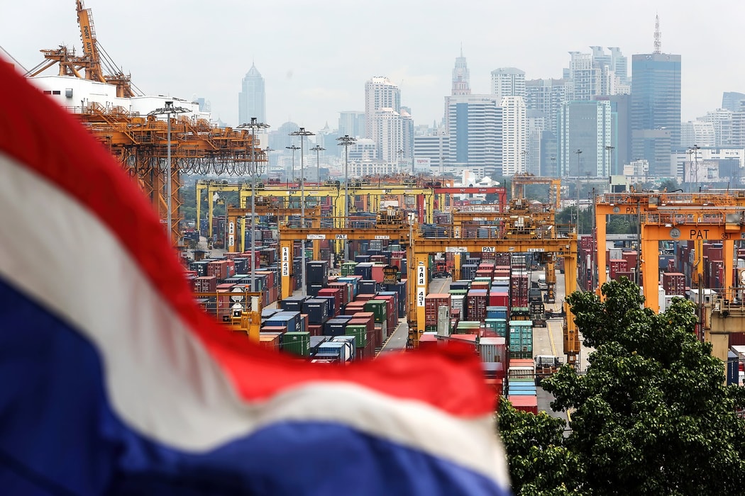 Quốc kỳ Thái Lan tung bay bên cạnh các container được xếp chồng gần cẩu giàn tại cảng Bangkok, với các tòa nhà cao tầng phía sau ở Bangkok, Thái Lan. Hình ảnh: Dario Pignatelli/Bloomberg.
